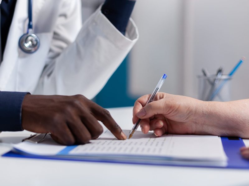 Close up of hands on desk at healthcare clinic Close up of hands on desk at healthcare clinic. Caucasian patient holding pen and signing document file for appointment checkup while african american doctor showing information